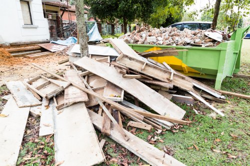 A pile of weathered wooden planks and broken timber is scattered across a grassy area adjacent to a construction or renovation site, with a large green disposal skip filled with mixed bricks, rubble, and debris in the background. The wooden materials vary in size and shape, some with painted or stained surfaces, and overlap each other in a disorganized manner. The scene includes a paved pathway and some leafy trees providing partial shade, indicative of an outdoor setting in a residential area, possibly within the vicinity of Tadworth. The overall context suggests removal and clearance of building waste, characteristic of rubbish removal services offered by companies like House Clearance Tadworth. The environment appears tidy apart from the debris, with natural light illuminating the scene and emphasizing the textures and colors of the discarded materials.