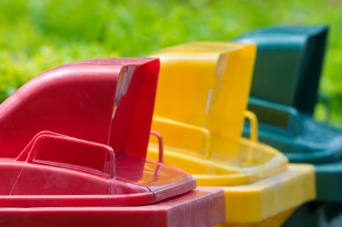 A row of four large plastic wheelie bins in red, yellow, and green colors are lined up on a pavement, with a blurred background of greenery and foliage. The bins are used for waste or rubbish collection, with the red bin positioned at the forefront, followed by the yellow and green bins. The lids are closed, and the bins are made of durable plastic with hinged lids and integrated handles for lifting and emptying. The image captures a typical scene outside a property or in a waste collection area, relevant to domestic rubbish removal services offered by companies such as House Clearance Tadworth, serving the Tadworth area within the postcode district. The natural outdoor environment and neatly arranged bins emphasize organized waste disposal and clearance activities.