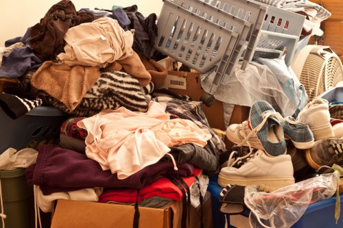A large pile of assorted household items and clothing materials arranged on a surface, including folded and crumpled garments in various colours such as beige, red, dark brown, and light pink, with textured fabrics like wool and cotton. Among the clothes, there are a few pairs of shoes, with one visible white sneaker and another covered in a plastic bag, placed on top of or beside the clothing. On the right side, a grey plastic laundry basket is tilted, filled with more textiles and possibly paper or cardboard, positioned next to a household appliance or part of the furniture. The background hints at an indoor setting, likely a room or storage area, with some parts of cardboard boxes and possibly curtains or other household items partially visible. This scene captures a typical cluttered environment often associated with rubbish or household clearing activities, relevant to domestic waste removal services offered by House Clearance Tadworth near the TA postcode area.