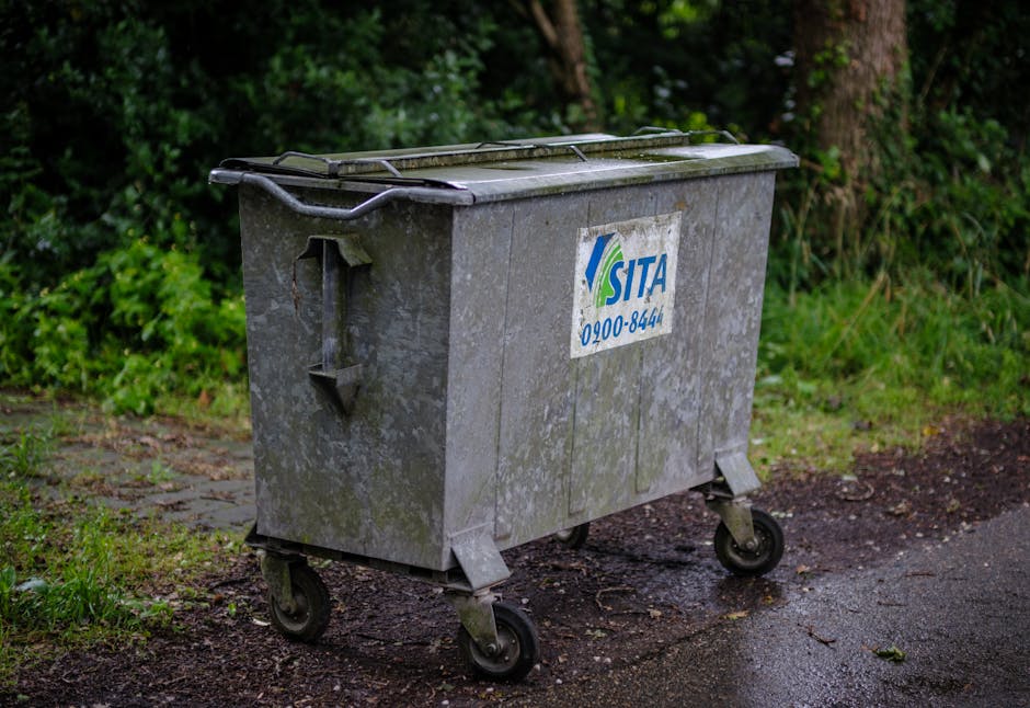 A large, rectangular metal rubbish bin with a hinged lid, situated outdoors on a paved surface adjacent to a grassy area with soil and scattered leaves. The bin is weathered with visible dirt, grime, 