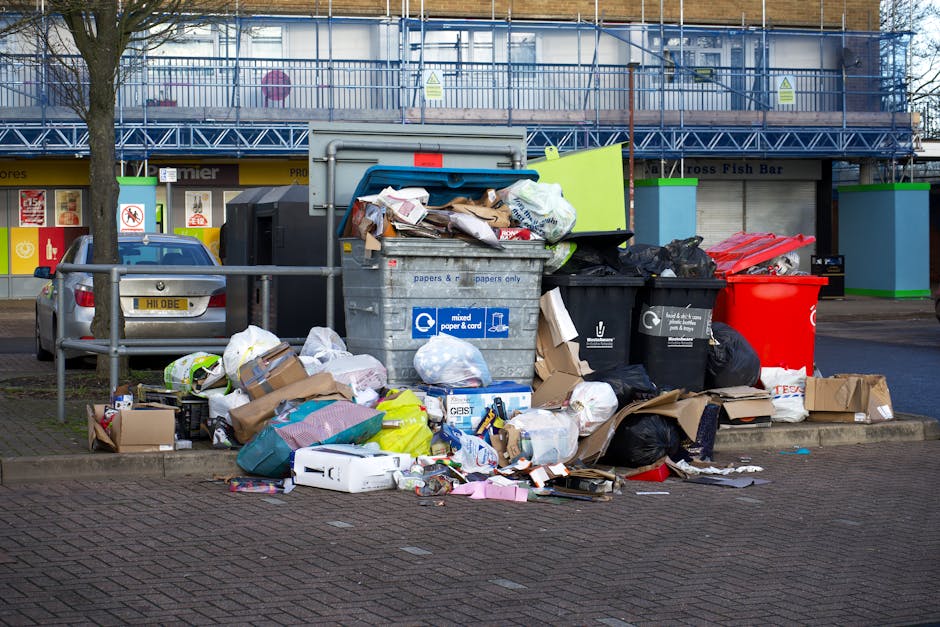 A large, rectangular metal rubbish bin with a hinged lid, situated outdoors on a paved surface adjacent to a grassy area with soil and scattered leaves. The bin is weathered with visible dirt, grime, 